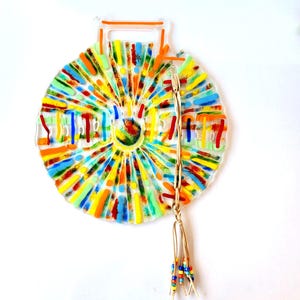 May include: A colorful glass suncatcher with a beaded tassel. The suncatcher is made of clear glass with a variety of colored lines and dots. The tassel is made of brown leather and has a variety of colored beads.