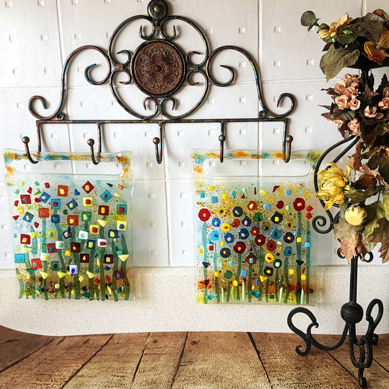 Op de afbeelding: Twee kleurrijke glazen panelen met abstracte bloemenmotieven hangen aan een metalen rek met haken. De panelen zijn gemaakt van helder glas met kleurrijke moza&iuml;ekstukken die een bloemenpatroon cre&euml;ren. De panelen hangen aan een metalen rek met haken.