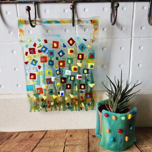 Op de afbeelding: Een kleurrijke glazen moza&iuml;ek wandhanger met een bloemenmotief. De moza&iuml;ek heeft vierkanten van rood, geel, blauw en groen glas. De hanger is bevestigd aan een witte muur met haken.