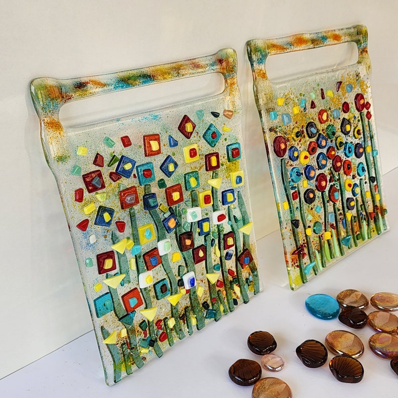 Op de afbeelding: Twee snijplanken van glas met een bloemenontwerp. De planken zijn gemaakt van helder glas met kleurrijke moza&iuml;ektegels in tinten rood, geel, blauw en groen. De tegels zijn gerangschikt in een patroon van bloemen en stengels.
