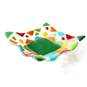 May include: A small, square glass dish with a wavy edge. The dish is decorated with a mosaic of colorful glass pieces in shades of red, orange, yellow, green, blue, and white. The center of the dish is a solid green square.