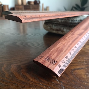 May include: Two wooden rulers with a reddish-brown grain finish. The rulers feature black printed markings for both inches and centimeters, with the metric scale clearly showing numbers up to 50 cm. A logo is stamped near the end of the ruler.