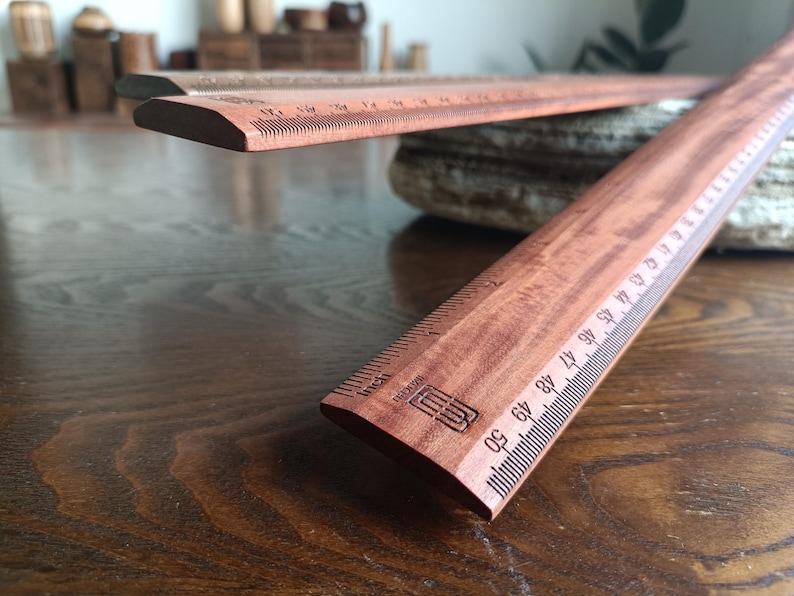 May include: Two wooden rulers with a reddish-brown grain finish. The rulers feature black printed markings for both inches and centimeters, with the metric scale clearly showing numbers up to 50 cm. A logo is stamped near the end of the ruler.