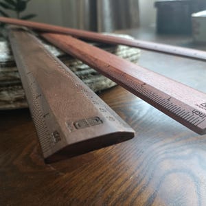May include: Two dark brown wooden rulers with beveled edges and printed measurement markings. Each ruler features a clear scale in inches along the side and a small logo stamped near the end. The wood grain is visible on the surface of both items.