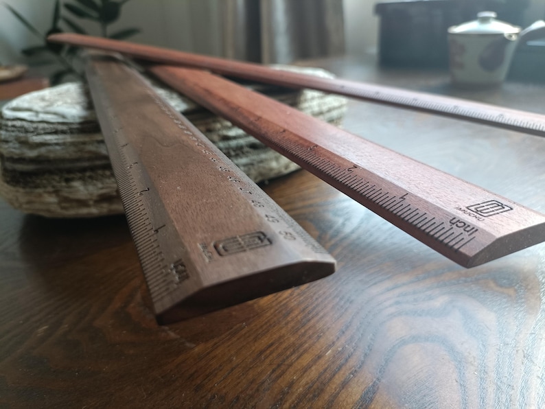 May include: Two dark brown wooden rulers with beveled edges and printed measurement markings. Each ruler features a clear scale in inches along the side and a small logo stamped near the end. The wood grain is visible on the surface of both items.