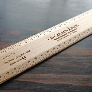 May include: A light-colored wooden architectural scale ruler featuring black printed markings and text. The ruler displays the text ON COMMON JOINERY, BESPOKE CABINETRY AND FURNITURE, by SCHROCKS of WALNUT CREEK, 1/2 in. = 1 ft., and contact information.