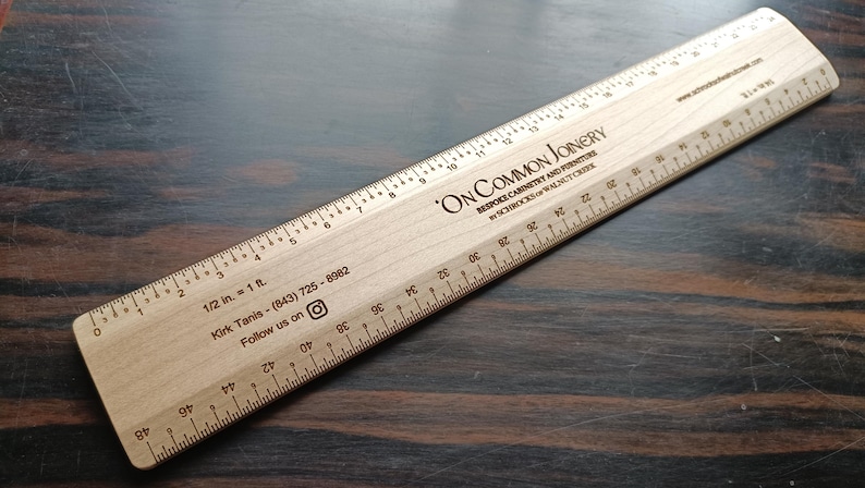 May include: A light-colored wooden architectural scale ruler featuring black printed markings and text. The ruler displays the text ON COMMON JOINERY, BESPOKE CABINETRY AND FURNITURE, by SCHROCKS of WALNUT CREEK, 1/2 in. = 1 ft., and contact information.