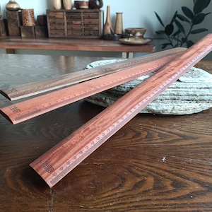 May include: Three wooden rulers with a reddish-brown wood grain finish. Each ruler features black printed metric markings along the edge, with the longest ruler clearly showing a scale up to 50 centimeters. The rulers have a flat, straight design.
