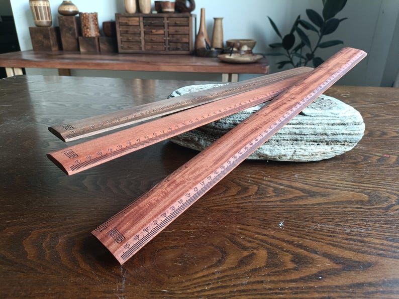 May include: Three wooden rulers with a reddish-brown wood grain finish. Each ruler features black printed metric markings along the edge, with the longest ruler clearly showing a scale up to 50 centimeters. The rulers have a flat, straight design.