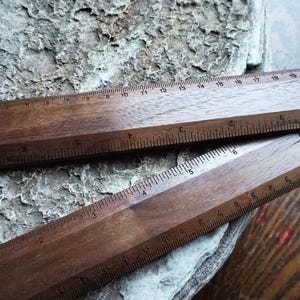 May include: Two dark brown wooden rulers with visible grain patterns. The top ruler features markings from 0 to 20 centimeters and 0 to 8 inches. The bottom ruler displays metric and imperial measurements with a small logo printed on the right side.