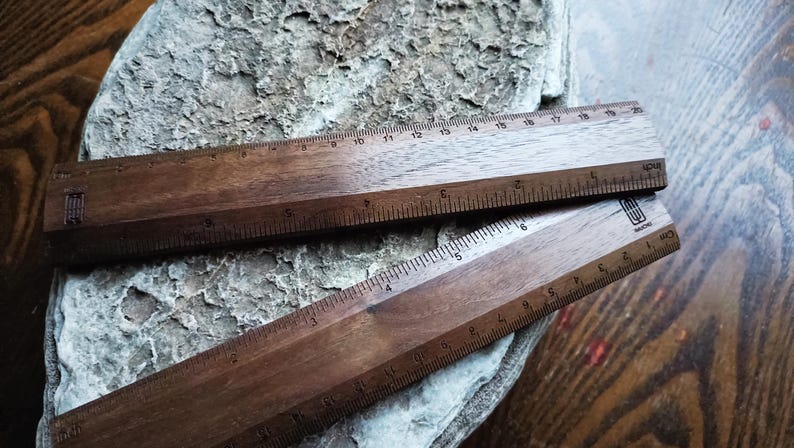May include: Two dark brown wooden rulers with visible grain patterns. The top ruler features markings from 0 to 20 centimeters and 0 to 8 inches. The bottom ruler displays metric and imperial measurements with a small logo printed on the right side.