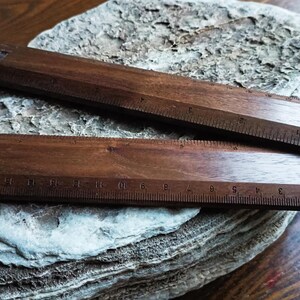 May include: Two dark brown wooden rulers with visible grain patterns. The top ruler features imperial markings in inches, while the bottom ruler displays metric markings in centimeters. A small black logo is printed on the bottom ruler's surface.