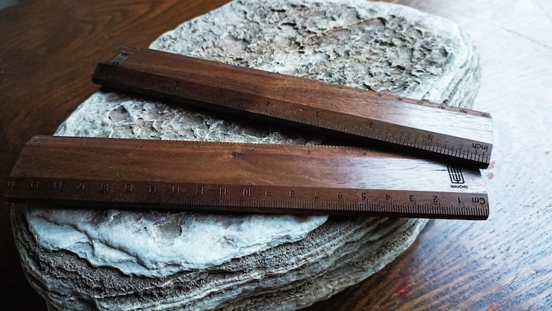 May include: Two dark brown wooden rulers with visible grain patterns. The top ruler features imperial markings in inches, while the bottom ruler displays metric markings in centimeters. A small black logo is printed on the bottom ruler's surface.