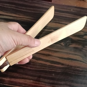 May include: A pair of light-colored wooden kitchen tongs featuring two flat, elongated arms connected at a hinge point with a dark brown accent piece. The tongs are constructed from smooth, natural wood with a visible grain pattern throughout the surface.