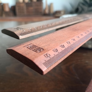 May include: Two wooden rulers with a reddish-brown finish, featuring clearly marked metric measurements up to 50 centimeters. Each ruler displays a small, rectangular logo stamped into the wood near the end, with precise black graduation lines.