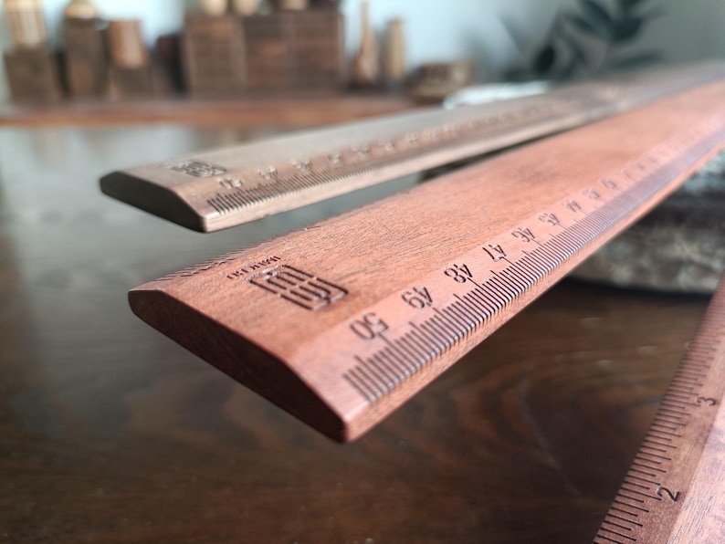 May include: Two wooden rulers with a reddish-brown finish, featuring clearly marked metric measurements up to 50 centimeters. Each ruler displays a small, rectangular logo stamped into the wood near the end, with precise black graduation lines.