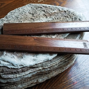 May include: Two dark brown wooden rulers with black measurement markings and a small black logo on the right side. The rulers feature a metric scale in centimeters, with the numbers 1 through 15 clearly visible along the bottom edge of each tool.