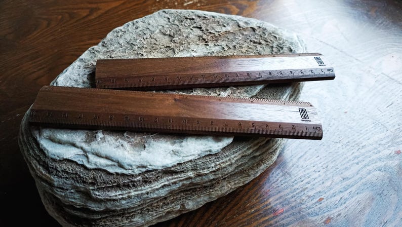 May include: Two dark brown wooden rulers with black measurement markings and a small black logo on the right side. The rulers feature a metric scale in centimeters, with the numbers 1 through 15 clearly visible along the bottom edge of each tool.