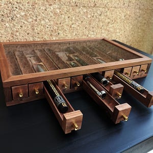 May include: A wooden box with a glass top and multiple drawers. The drawers are open and reveal black pens inside. The box is made of dark wood with brass accents.