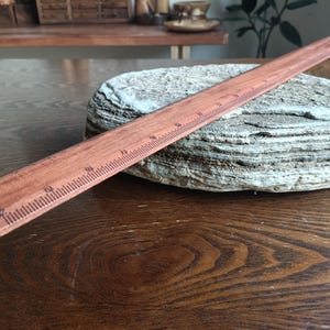 May include: A straight wooden ruler with a reddish-brown finish and dark brown measurement markings. The ruler features clear numerical increments along its edge, displaying a standard scale for measuring length in inches and fractions of an inch.