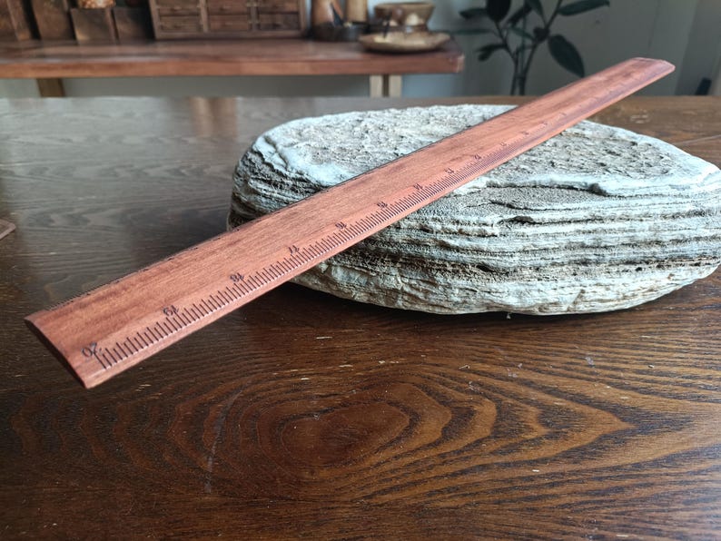 May include: A straight wooden ruler with a reddish-brown finish and dark brown measurement markings. The ruler features clear numerical increments along its edge, displaying a standard scale for measuring length in inches and fractions of an inch.