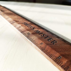 May include: A wooden ruler with a dark brown finish and a visible wood grain pattern. The ruler features black engraved measurement markings and the text "SWISS MISTER" clearly etched into the surface, showing numbers up to 31 inches.