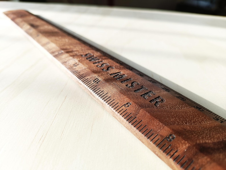May include: A wooden ruler with a dark brown finish and a visible wood grain pattern. The ruler features black engraved measurement markings and the text "SWISS MISTER" clearly etched into the surface, showing numbers up to 31 inches.