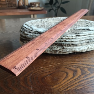 May include: A straight wooden ruler with a reddish-brown finish and dark engraved markings. The ruler features clear numerical increments, with the number 20 visible at the front edge, indicating measurements in inches along its length.