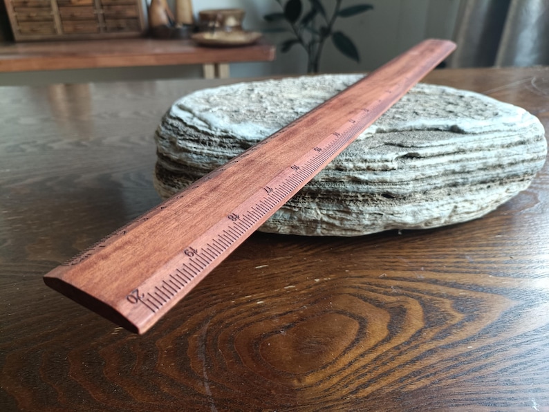 May include: A straight wooden ruler with a reddish-brown finish and dark engraved markings. The ruler features clear numerical increments, with the number 20 visible at the front edge, indicating measurements in inches along its length.