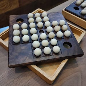 May include: A wooden game board with a walnut finish and white marbles. The board has a grid pattern with holes in the corners. The board is open and the marbles are visible.