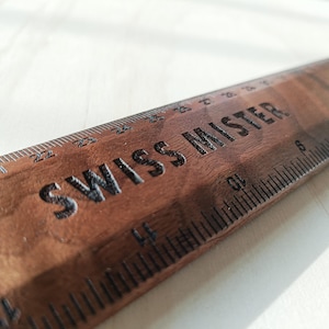 May include: A dark brown wooden ruler featuring the engraved text SWISS MISTER. The ruler displays clear measurement markings in inches along the edge, with visible numbers ranging from 12 to 27 inches etched into the wood surface.