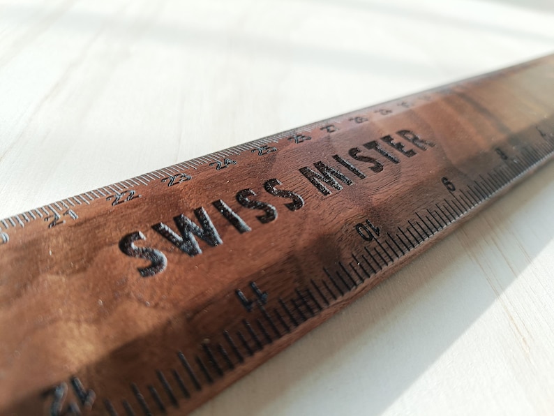 May include: A dark brown wooden ruler featuring the engraved text SWISS MISTER. The ruler displays clear measurement markings in inches along the edge, with visible numbers ranging from 12 to 27 inches etched into the wood surface.