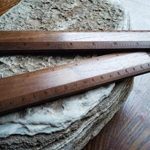 May include: Two dark brown wooden rulers with visible grain patterns. The top ruler features imperial markings in inches, while the bottom ruler displays metric markings in centimeters. Both rulers have a small logo printed on the right side.