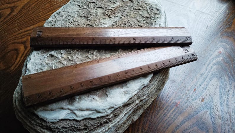May include: Two dark brown wooden rulers with visible grain patterns. The top ruler features imperial markings in inches, while the bottom ruler displays metric markings in centimeters. Both rulers have a small logo printed on the right side.