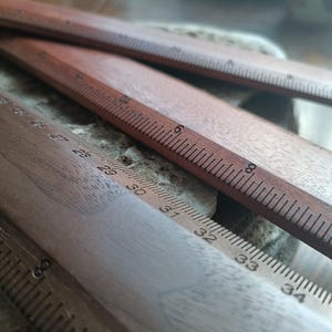 May include: Three wooden rulers with visible measurement markings. One ruler displays imperial units with numbers up to 8 inches, while another shows metric units with numbers ranging from 26 to 35 centimeters, clearly printed on the wood surface.
