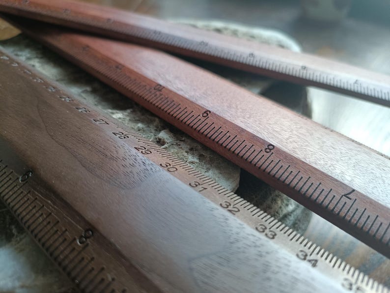 May include: Three wooden rulers with visible measurement markings. One ruler displays imperial units with numbers up to 8 inches, while another shows metric units with numbers ranging from 26 to 35 centimeters, clearly printed on the wood surface.