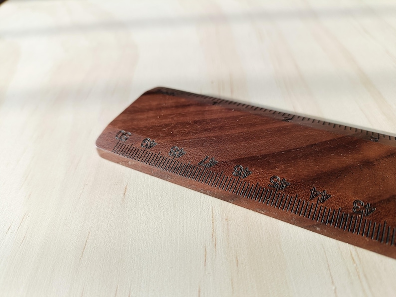 May include: A dark brown wooden ruler featuring clearly etched black measurement markings. The ruler displays numerical increments starting from 43 and continuing upward, with a visible grain pattern throughout the wood surface.