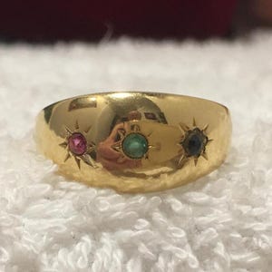 Könnte beinhalten: Ein goldener Ring mit drei Edelsteinen: einem Rubin, einem Smaragd und einem Saphir. Jeder Stein ist in einem sternförmigen Muster gefasst. Der Ring hat eine glatte, polierte Oberfläche und eine abgerundete Form.