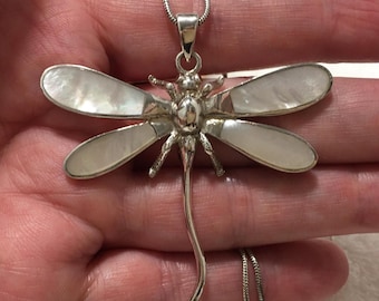 May include: Silver dragonfly pendant necklace with mother-of-pearl inlay wings. The dragonfly pendant is detailed with a textured body and delicate legs. The necklace chain is a thin, silver snake chain.