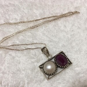 Op de afbeelding: Een zilveren ketting met een rechthoekige hanger. De hanger heeft een witte parel en een rode robijn. De ketting is delicaat en de hanger zit in een zilveren frame. Sieraden.