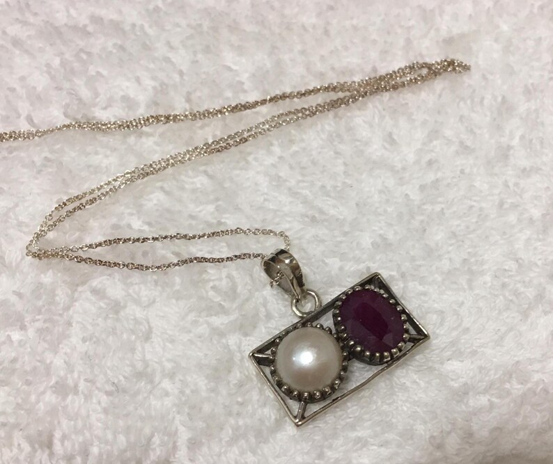 Op de afbeelding: Een zilveren ketting met een rechthoekige hanger. De hanger heeft een witte parel en een rode robijn. De ketting is delicaat en de hanger zit in een zilveren frame. Sieraden.