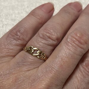 May include: A gold-colored ring with a chain link design. The ring is worn on a finger and has a polished finish. The ring is a piece of jewelry.