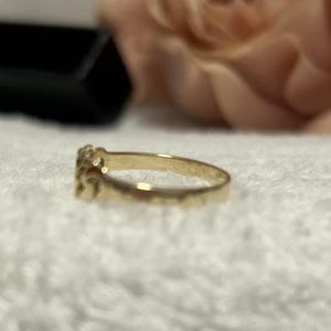 May include: A gold ring with a decorative design. The ring is set against a white, textured surface, with a blurred background that includes a black box and a pinkish-beige flower.