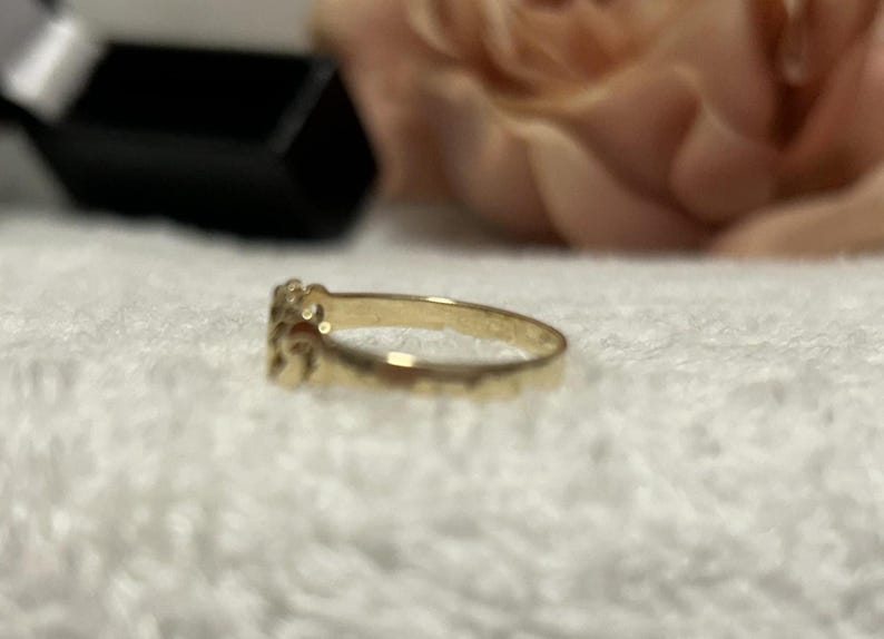 May include: A gold ring with a decorative design. The ring is set against a white, textured surface, with a blurred background that includes a black box and a pinkish-beige flower.