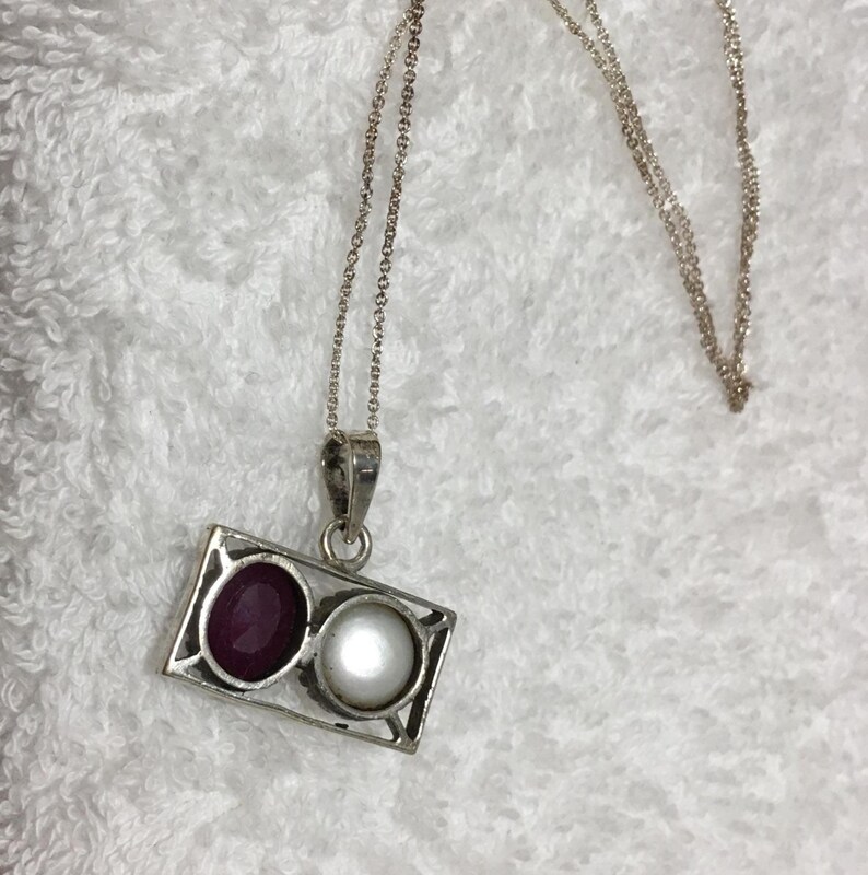 Op de afbeelding: Zilveren hangerketting met een rechthoekig frame met een donkerrode edelsteen en een witte parel. De hanger hangt aan een fijne zilveren ketting en biedt een klassieke en elegante uitstraling. De ketting wordt getoond op een witte, getextureerde achtergrond.