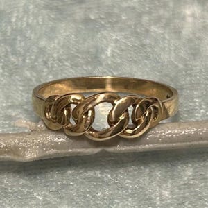May include: A gold-colored ring with a chain link design. The ring features three interconnected chain links across the top, creating a bold and stylish look. The ring is set against a light blue background.