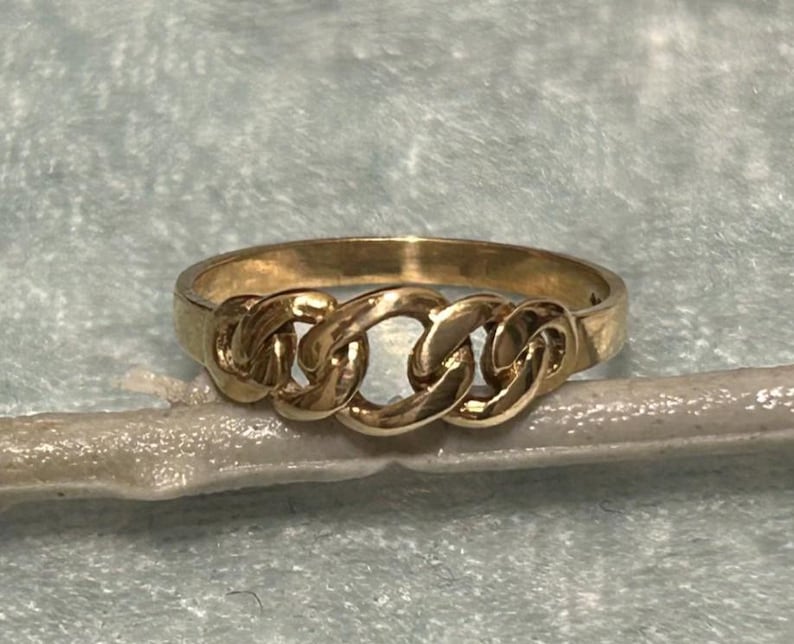 May include: A gold-colored ring with a chain link design. The ring features three interconnected chain links across the top, creating a bold and stylish look. The ring is set against a light blue background.