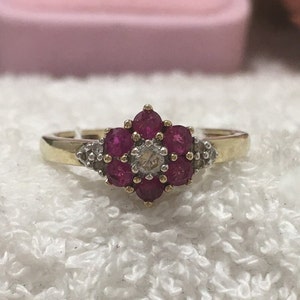 May include: A gold ring with a flower-shaped cluster of red gemstones and a central diamond. The ring is displayed on a white, textured background, highlighting the jewellery's design and craftsmanship.