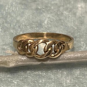 May include: A gold-colored ring with a chain link design. The ring features three interconnected chain links across the top, with a smooth band. The ring is set against a light blue background.