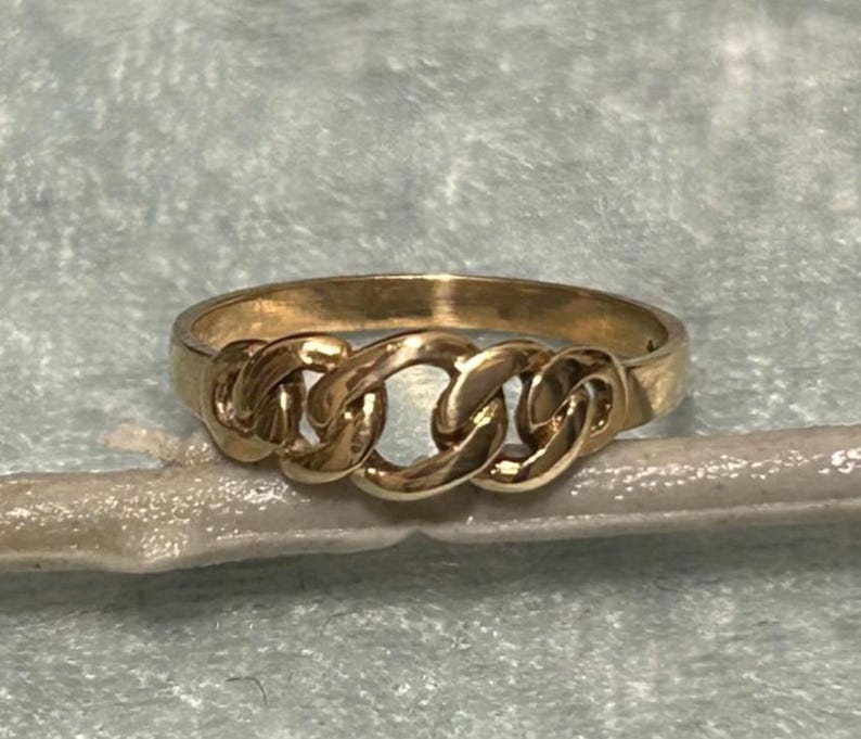 May include: A gold-colored ring with a chain link design. The ring features three interconnected chain links across the top, with a smooth band. The ring is set against a light blue background.